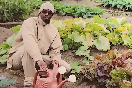 A photo of L crouching surrounded by vegetables he grew, his hand resting on an orange watering can.