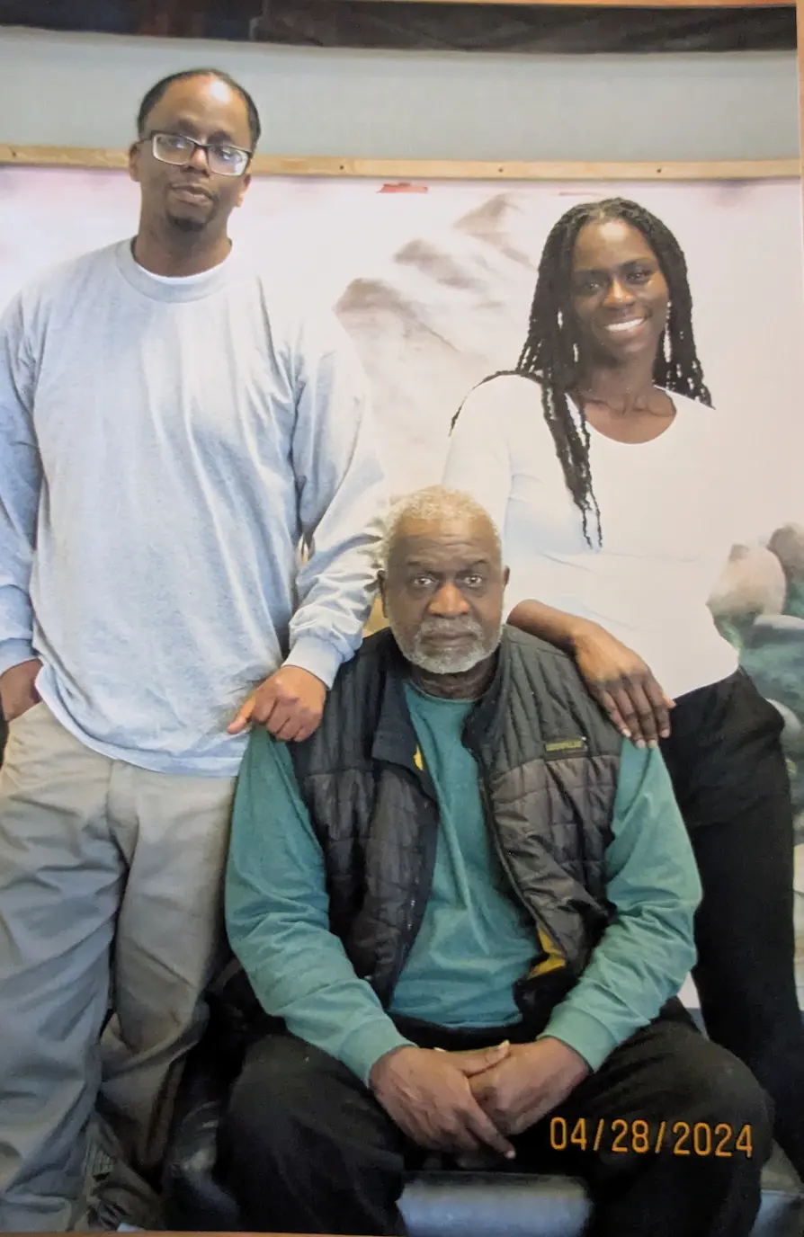 A photo of L and a woman standing on either side of an older man with grey hair sitting in a chair.
